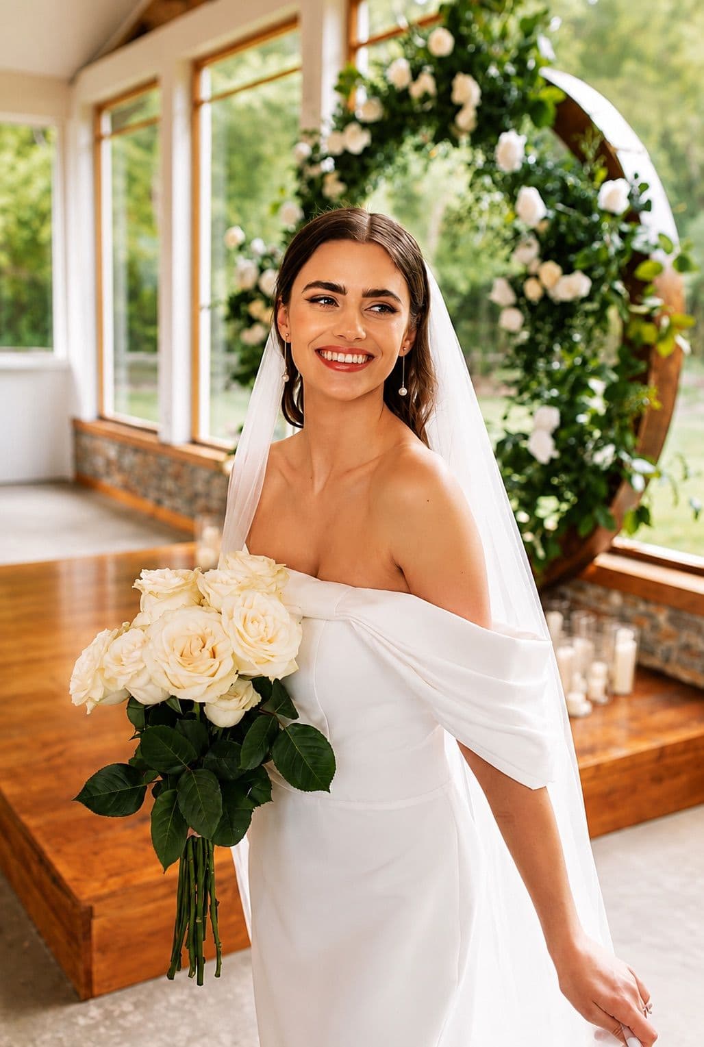 A smiling bride in an off-the-shoulder gown holding a bouquet of white roses.
