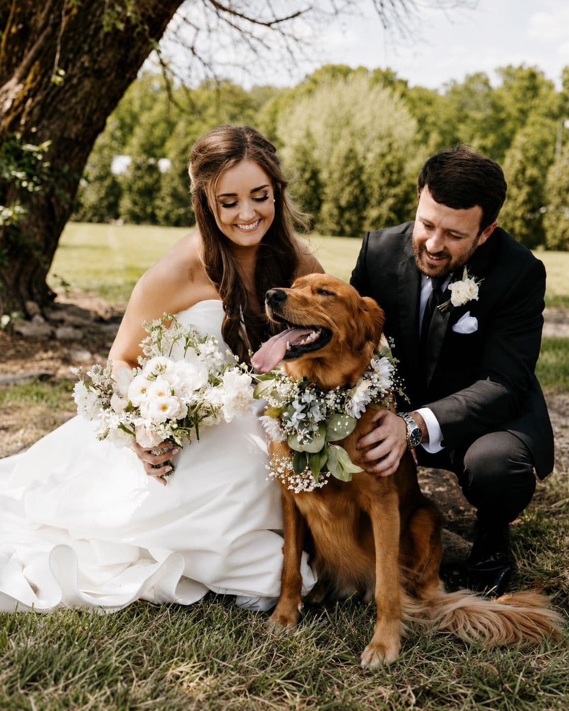 A bride and groom kneeling on the grass beside their golden retriever wearing a floral collar.