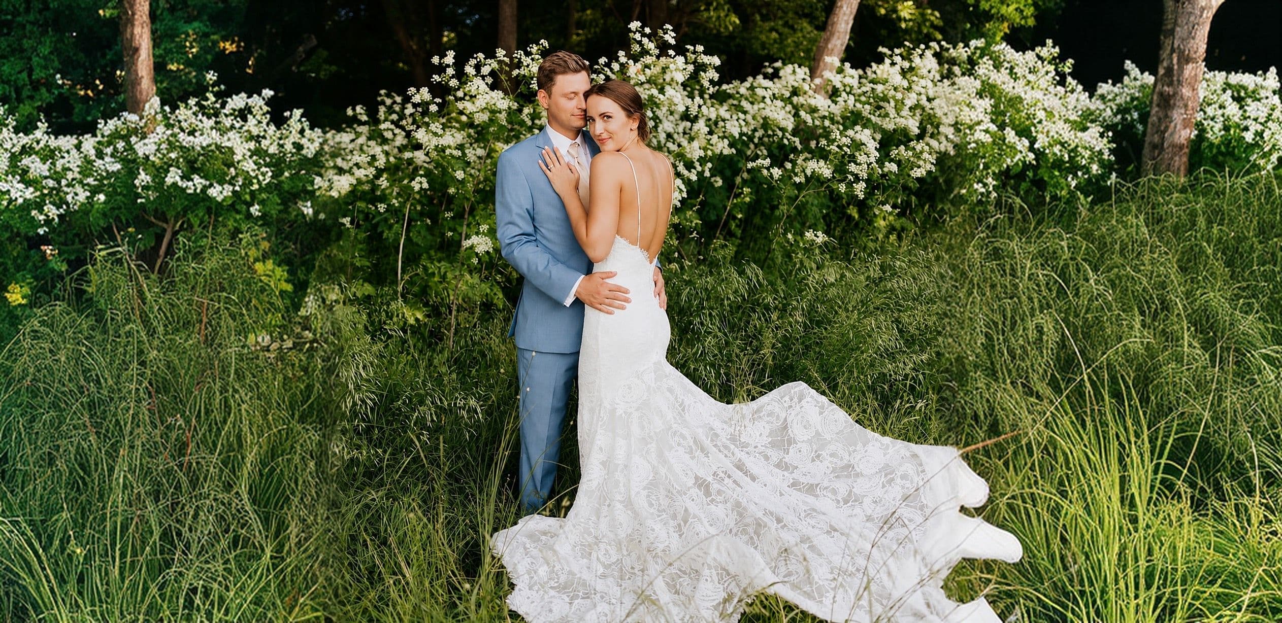 A bride and groom embracing in a meadow of white wildflowers, her lace train spread across the grass.
