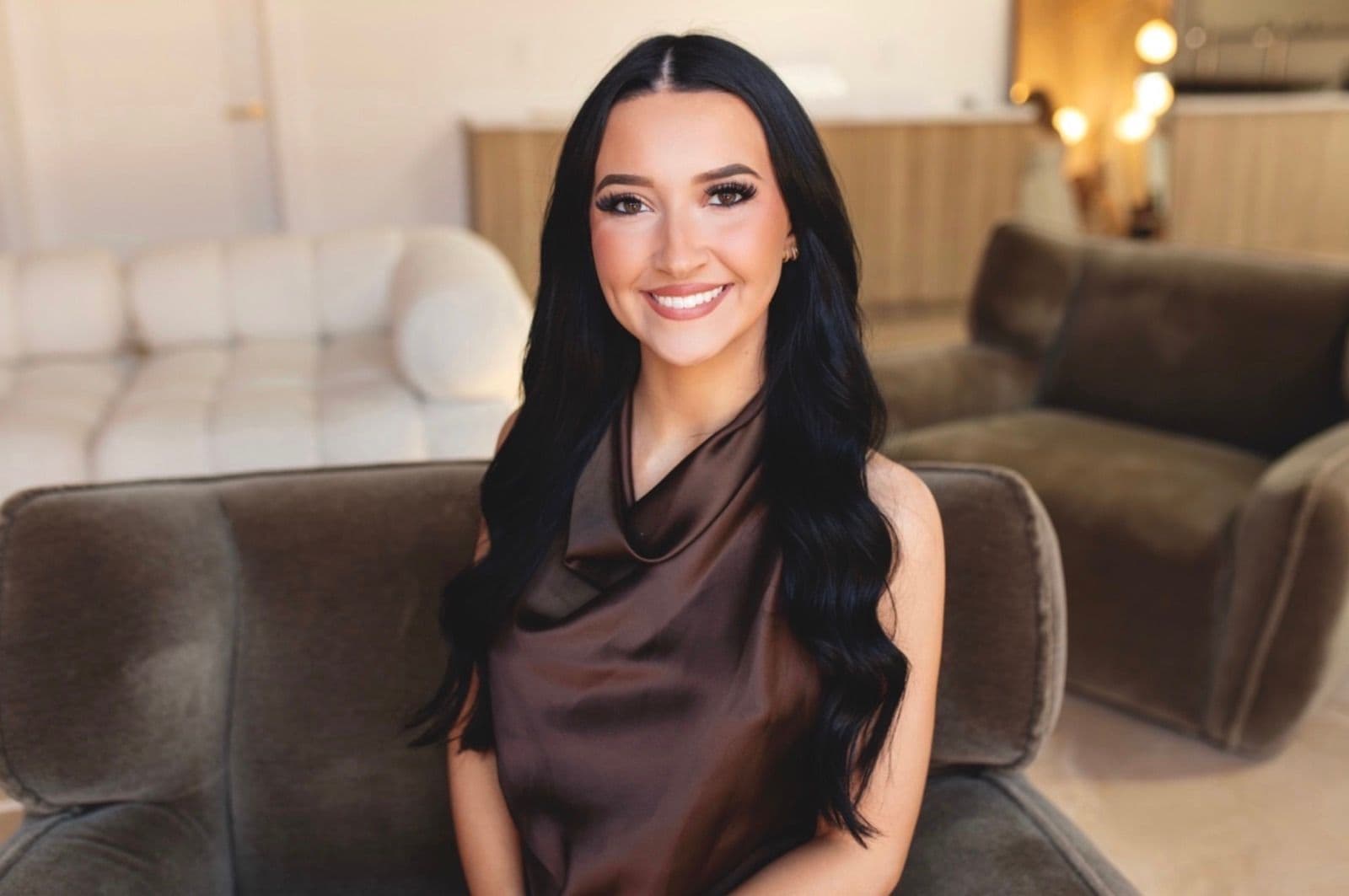 Chesney Pope, smiling, with long dark hair, seated in a velvet armchair.