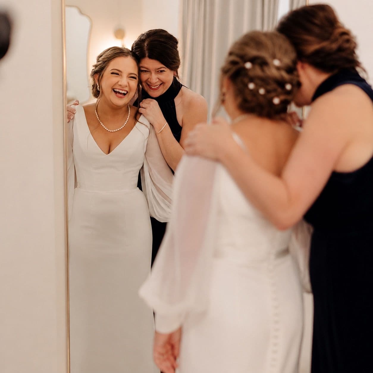 A laughing bride hugged by a friend in front of a mirror while getting ready.